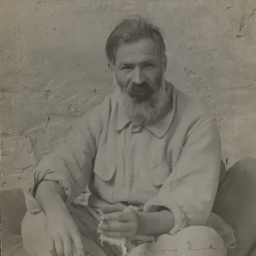 Black-and-white portrait photograph of a bearded man in light-colored clothing, seated and gazing directly at the camera.