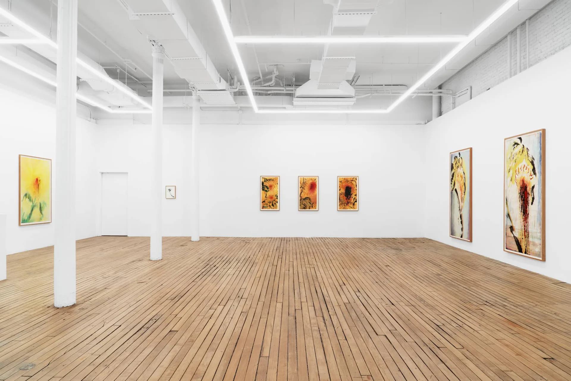 Bright, minimalist gallery interior with white walls, wooden floors, and support columns; colorful floral paintings displayed on walls.