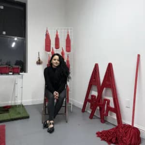 Red fabric and paper installation with tall cone-shaped forms, person seated beside sculptural elements in gallery space.