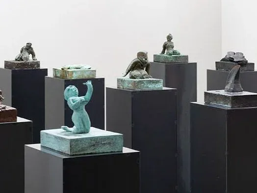 Gallery installation view with black pedestals supporting patinated bronze sculptural figures, white walls, gallery lighting.