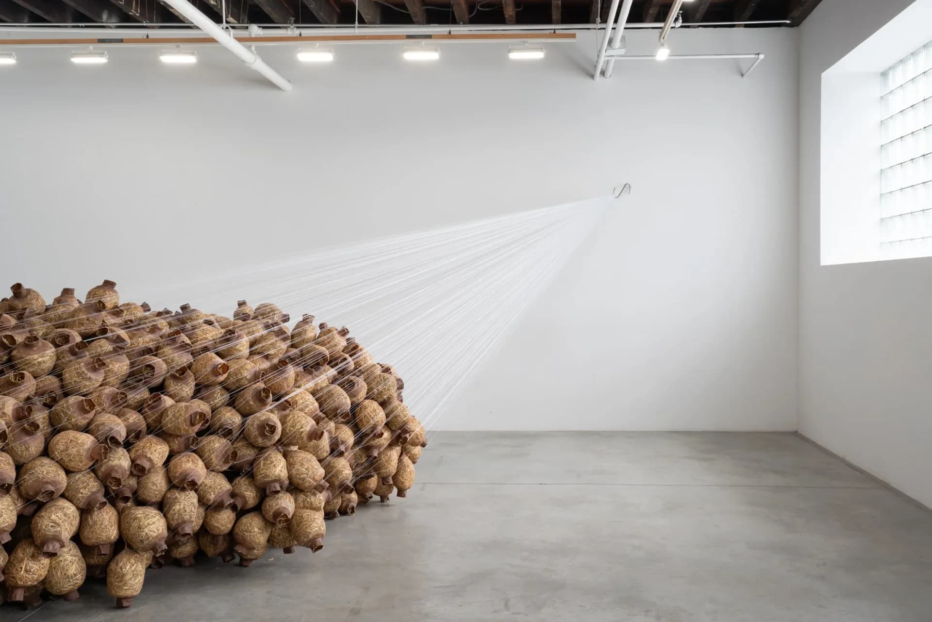 Large sculptural installation of stacked brown woven spherical forms heaped against a white gallery wall with natural light.