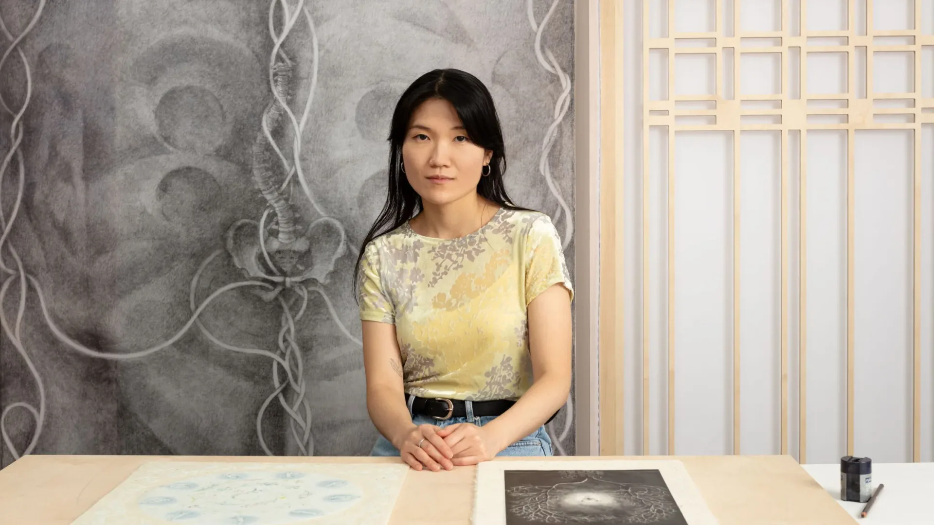 Woman with long black hair wearing a pale yellow short-sleeved top and black belt, seated at a desk.