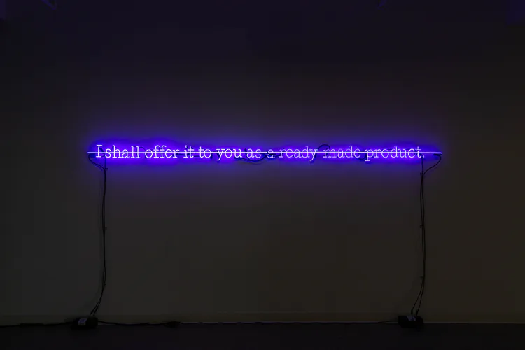 Blue neon text installation reading 'i shall offer it to you as a ready made product' mounted on dark wall.