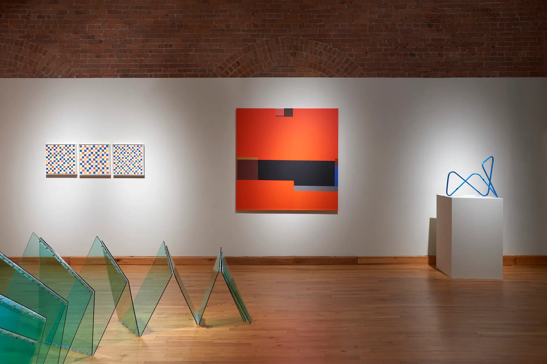 Gallery space with wooden floor displaying geometric glass sculptures, patterned wall art, a red and black painting, and a sculptural piece on pedestal.