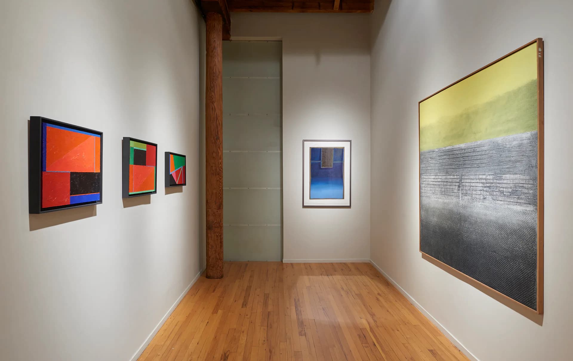 Narrow gallery corridor with wooden flooring and exposed beam, displaying abstract paintings in orange, black, and blue tones on white walls.