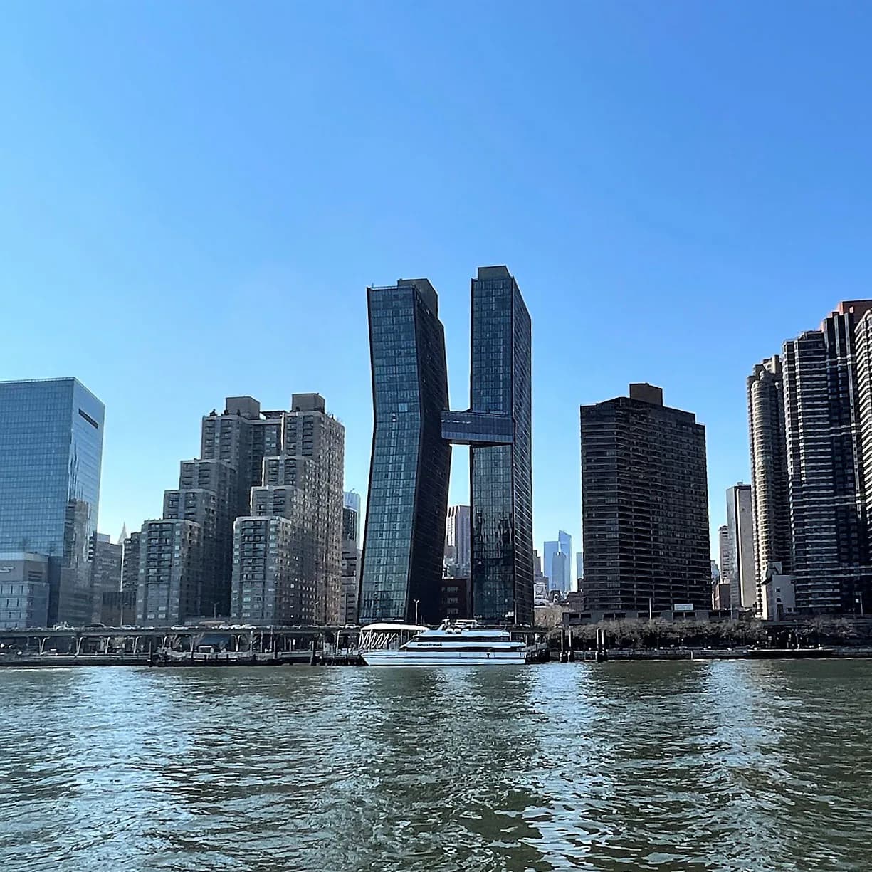Photograph of Manhattan skyline viewed from East River with contemporary skyscrapers and clear blue sky.