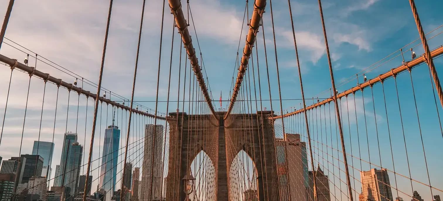Brooklyn Bridge with Gothic stone towers and steel cables framing Manhattan skyline under blue sky with white clouds.
