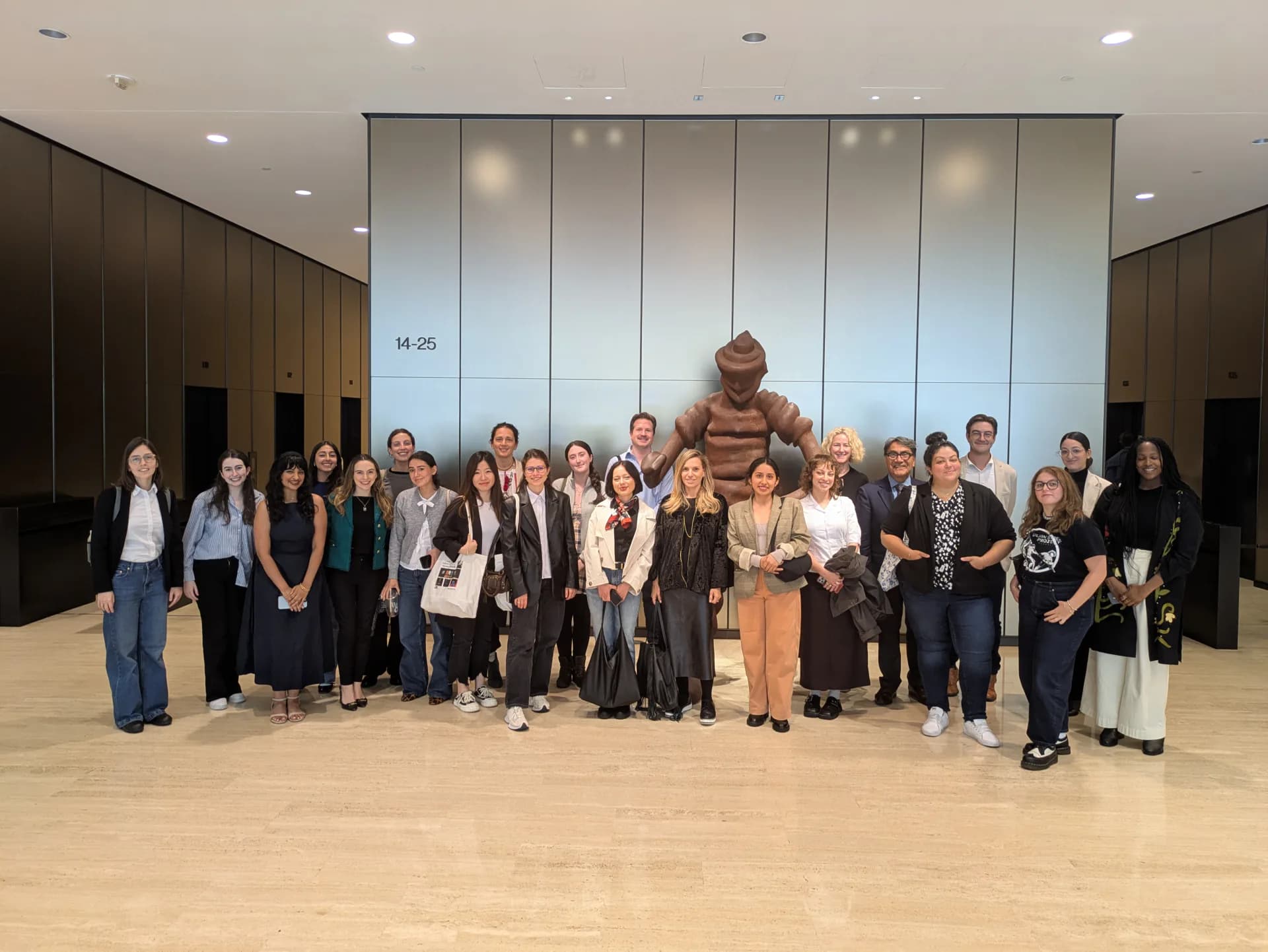 Large group of approximately 30 people standing in a modern gallery interior with glass partition walls and wooden flooring.