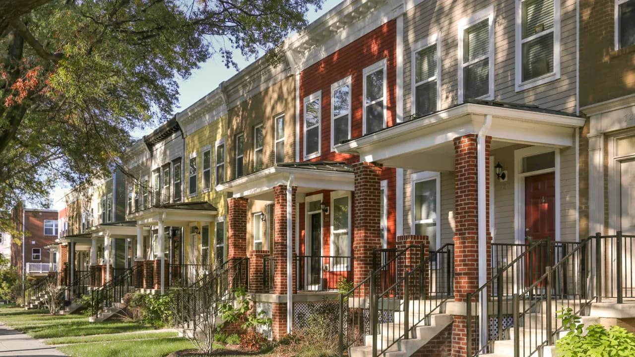 Tree-lined street of historic rowhouses with red brick, pale facades, wrought-iron railings, storefronts, and manicured front yards.