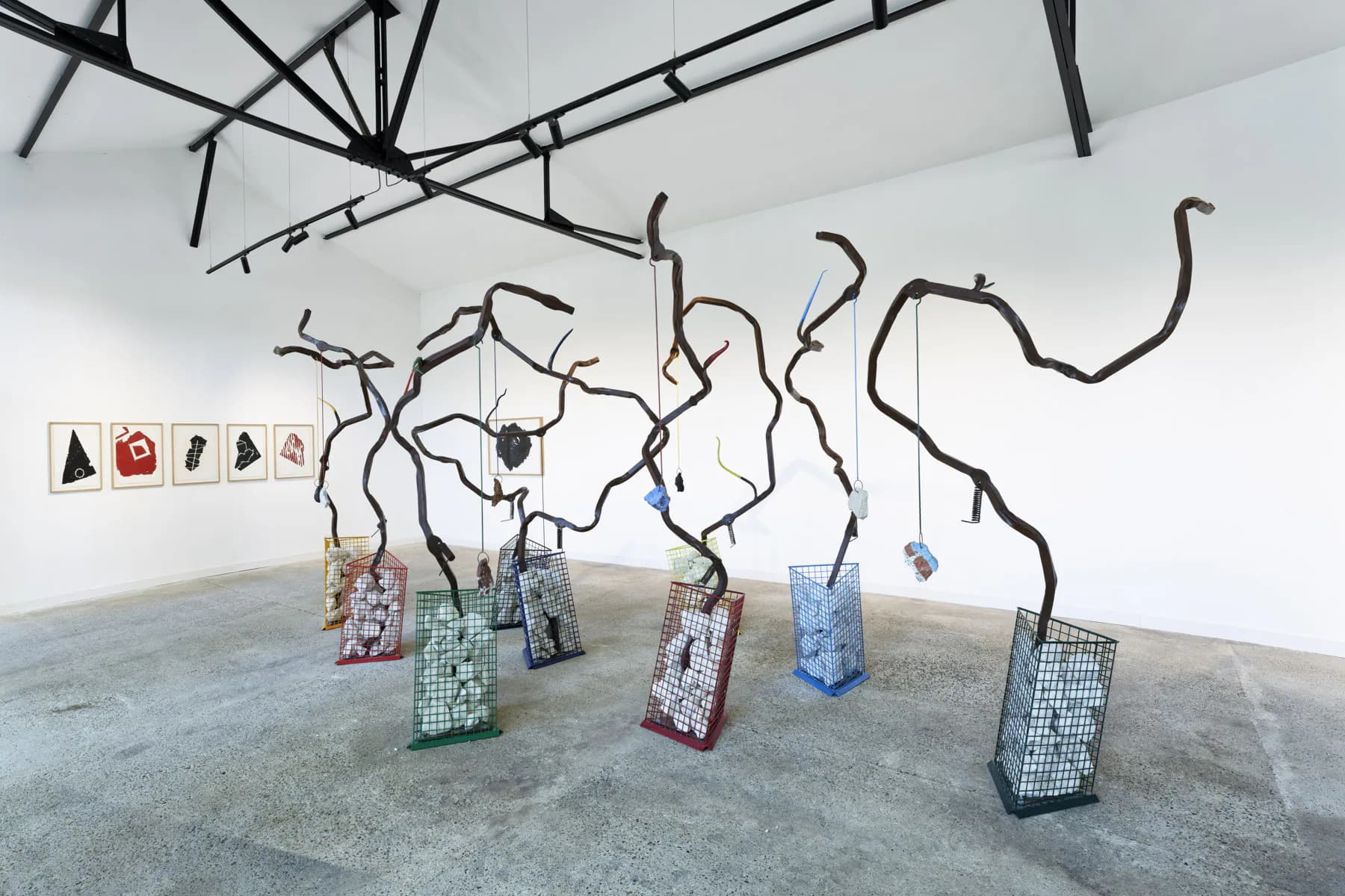 Installation with twisted black metal branches extending from ceiling and floor, supporting colorful patterned fabric baskets and hanging objects.