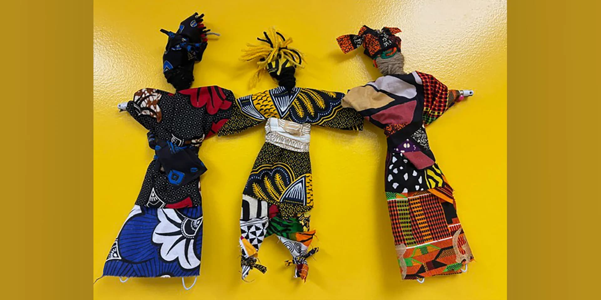Three fabric dolls with black heads and colorful patterned cloth bodies in blue, yellow, and red African-inspired prints arranged on bright yellow background.