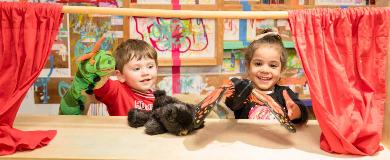 Two young children perform with a puppet in a colorful puppet theater with red curtains at Children's Museum of Manhattan.