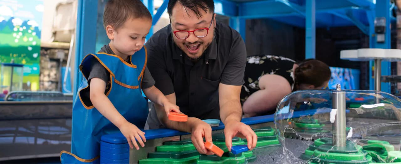 An adult and child collaborate on a green building project at an interactive table in a blue-walled museum space.