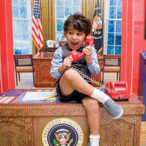 A child sits on the Resolute Desk in a replica Oval Office space, holding a red telephone prop.