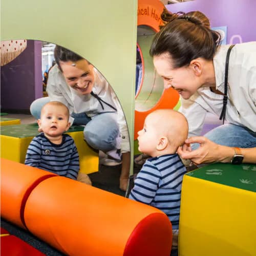 Two adults guide toddlers through a colorful play tunnel with orange and yellow elements in an interactive museum exhibit.