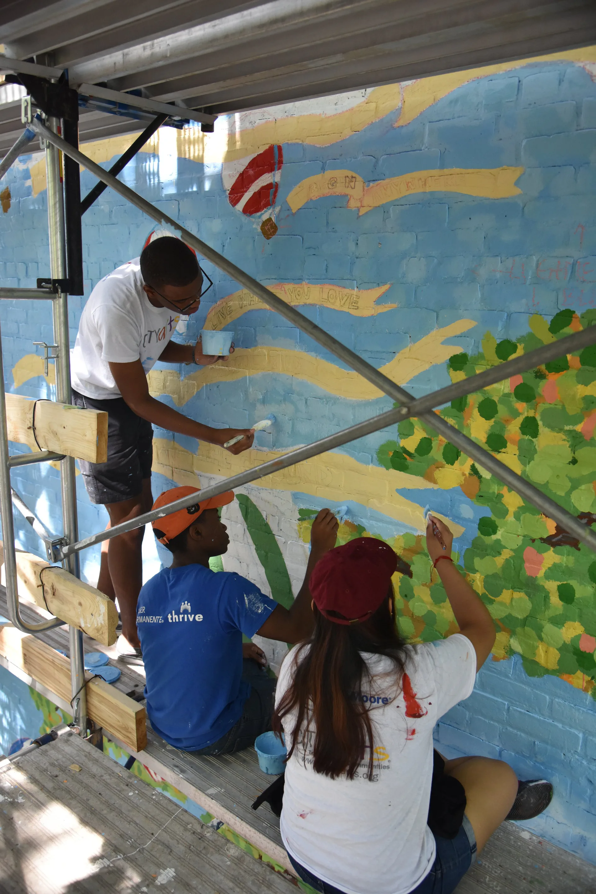 Three people painting a large mural on a blue wall with yellow geometric shapes and green foliage details under scaffolding.