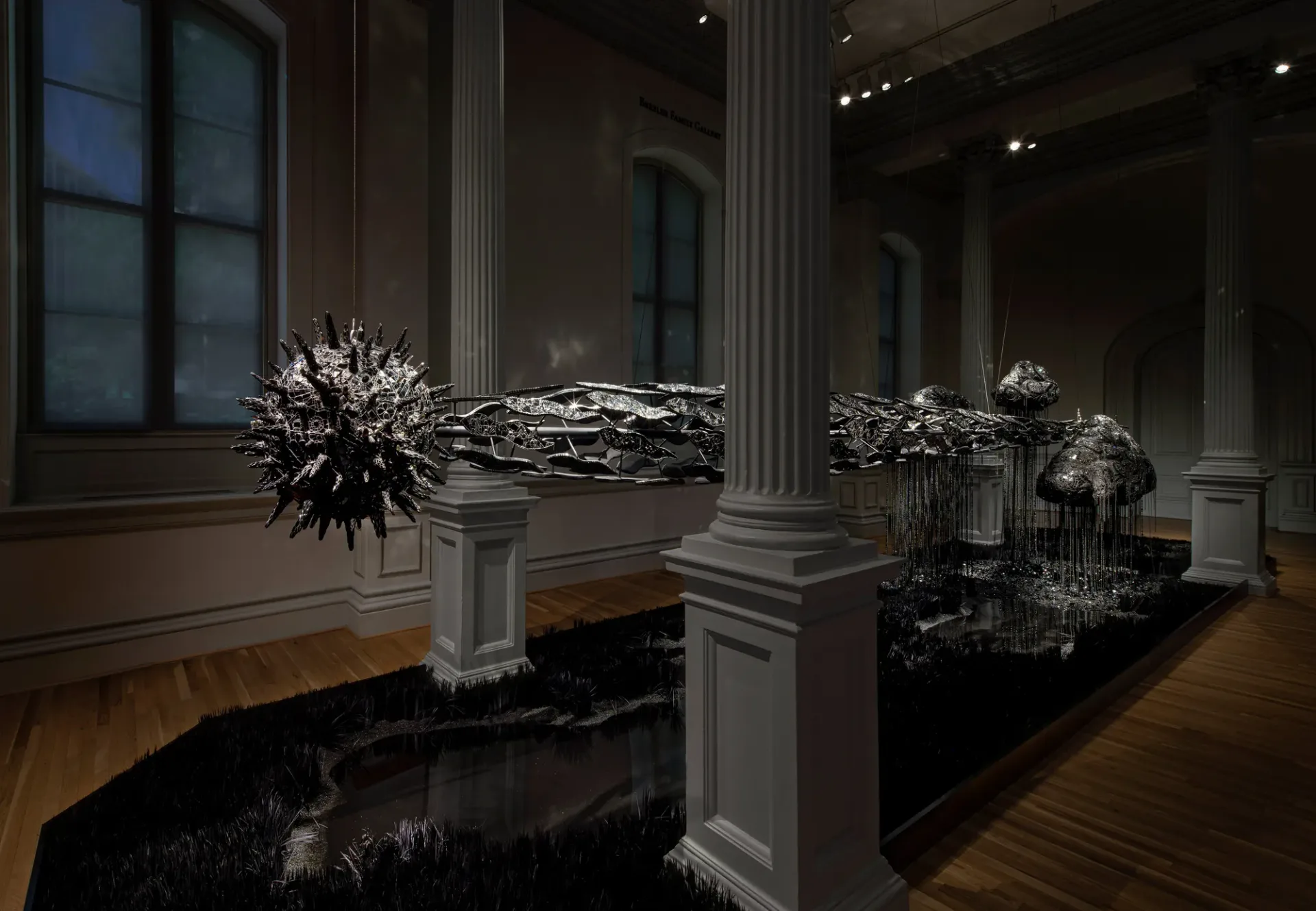 Photograph of a gallery interior with classical columns and dark sculptural installations featuring spiky, urchin-like forms displayed on white pedestals on a wooden floor.