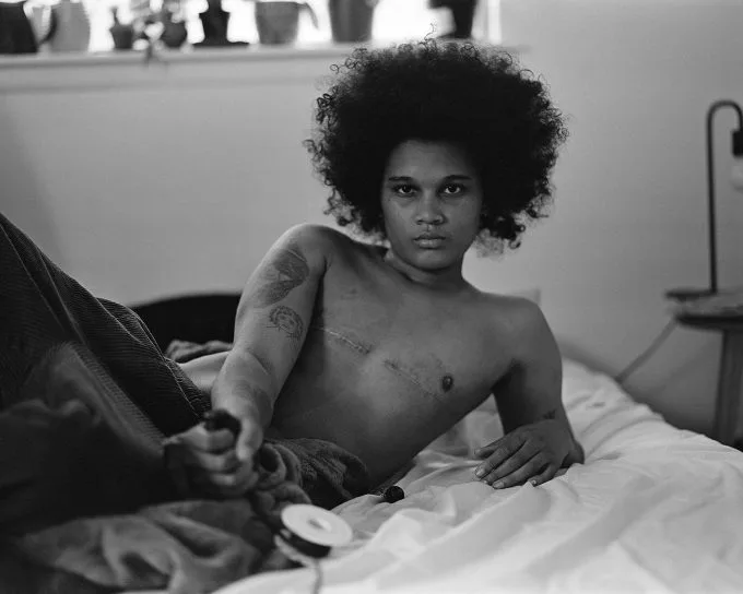 Black and white photograph of a young child with natural hair, shirtless on white bedding, gazing at camera.