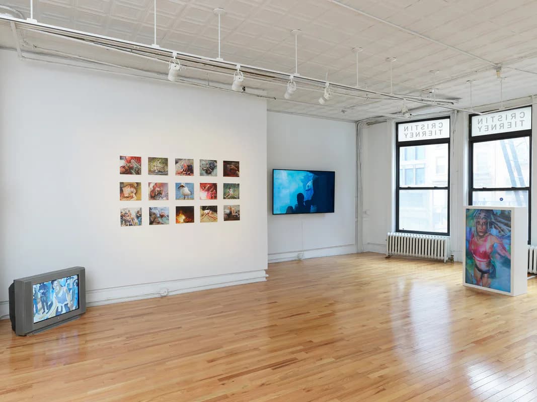 Bright gallery with grid of colorful small artworks on left wall, blue monitor, wooden floor, and mixed media displayed.