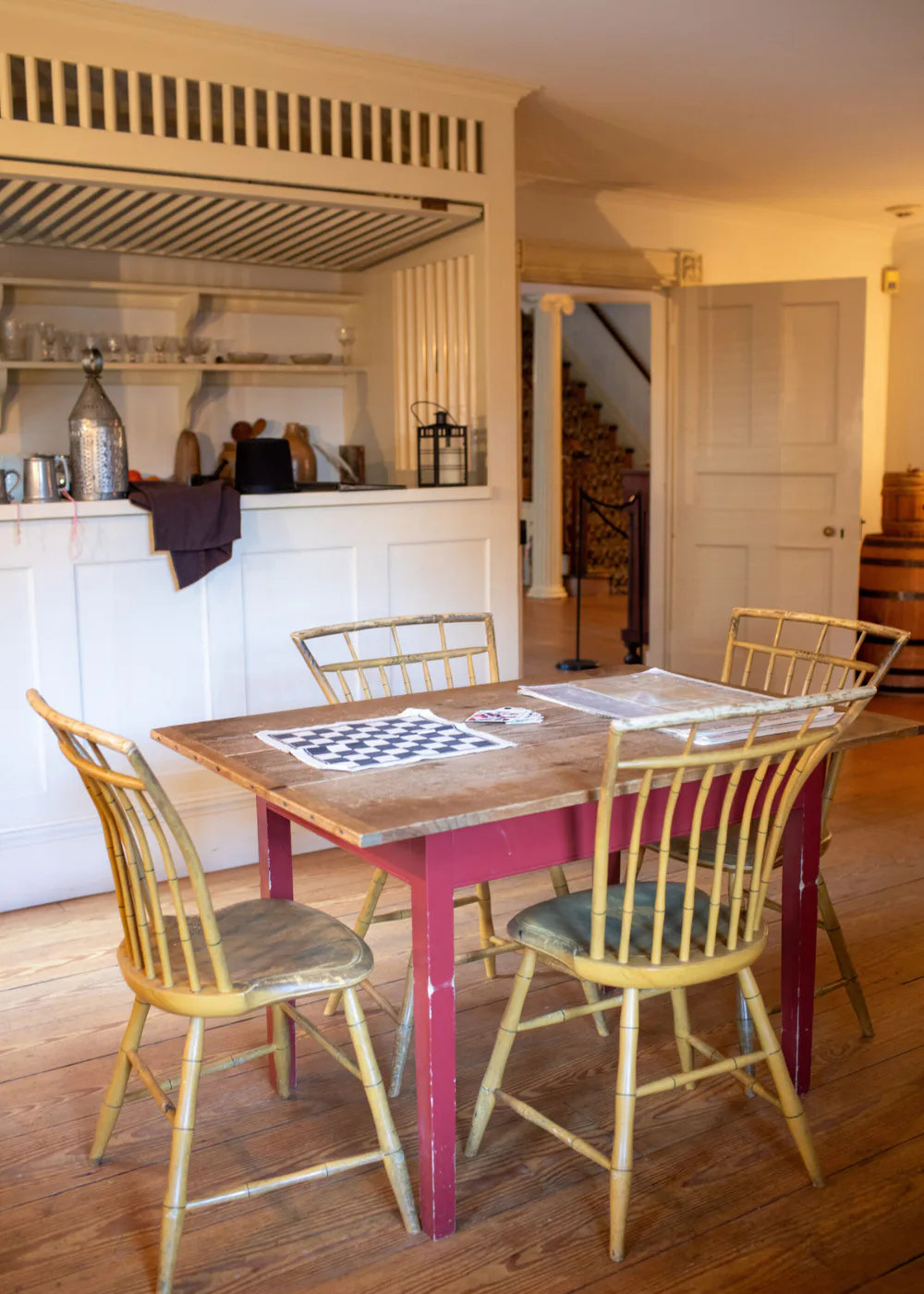 Interior photograph of a modern kitchenette and dining area with yellow spindle-back chairs, a wooden table with pink base, white cabinetry, and warm lighting.