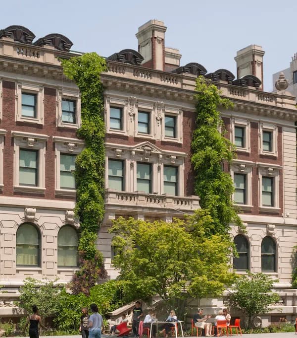 Exterior of a red-brick institutional building with ivy climbing the facade, people gathered at outdoor seating below.