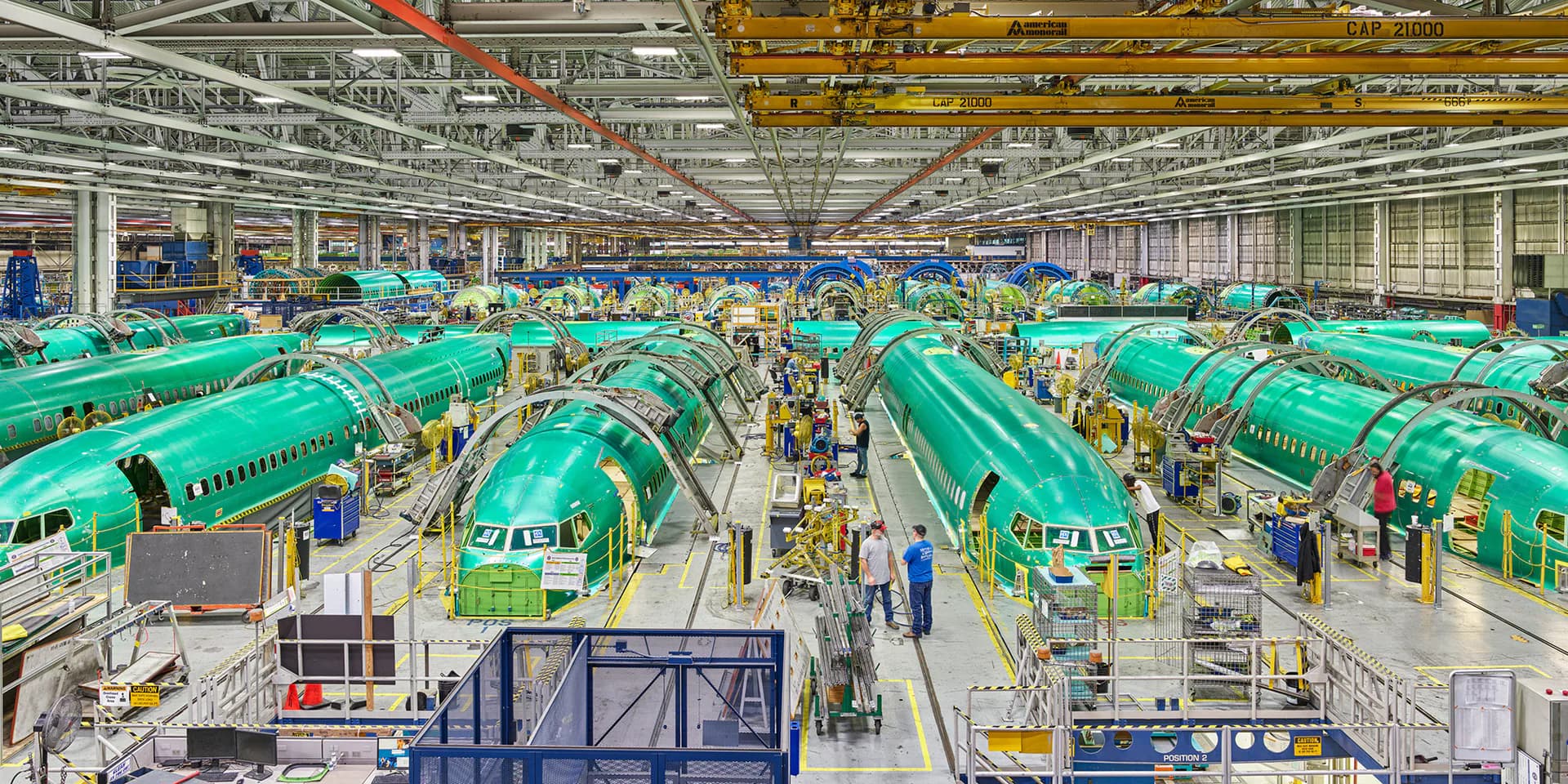 Industrial factory floor with rows of turquoise aircraft fuselages under construction, bright overhead lighting, workers visible throughout.
