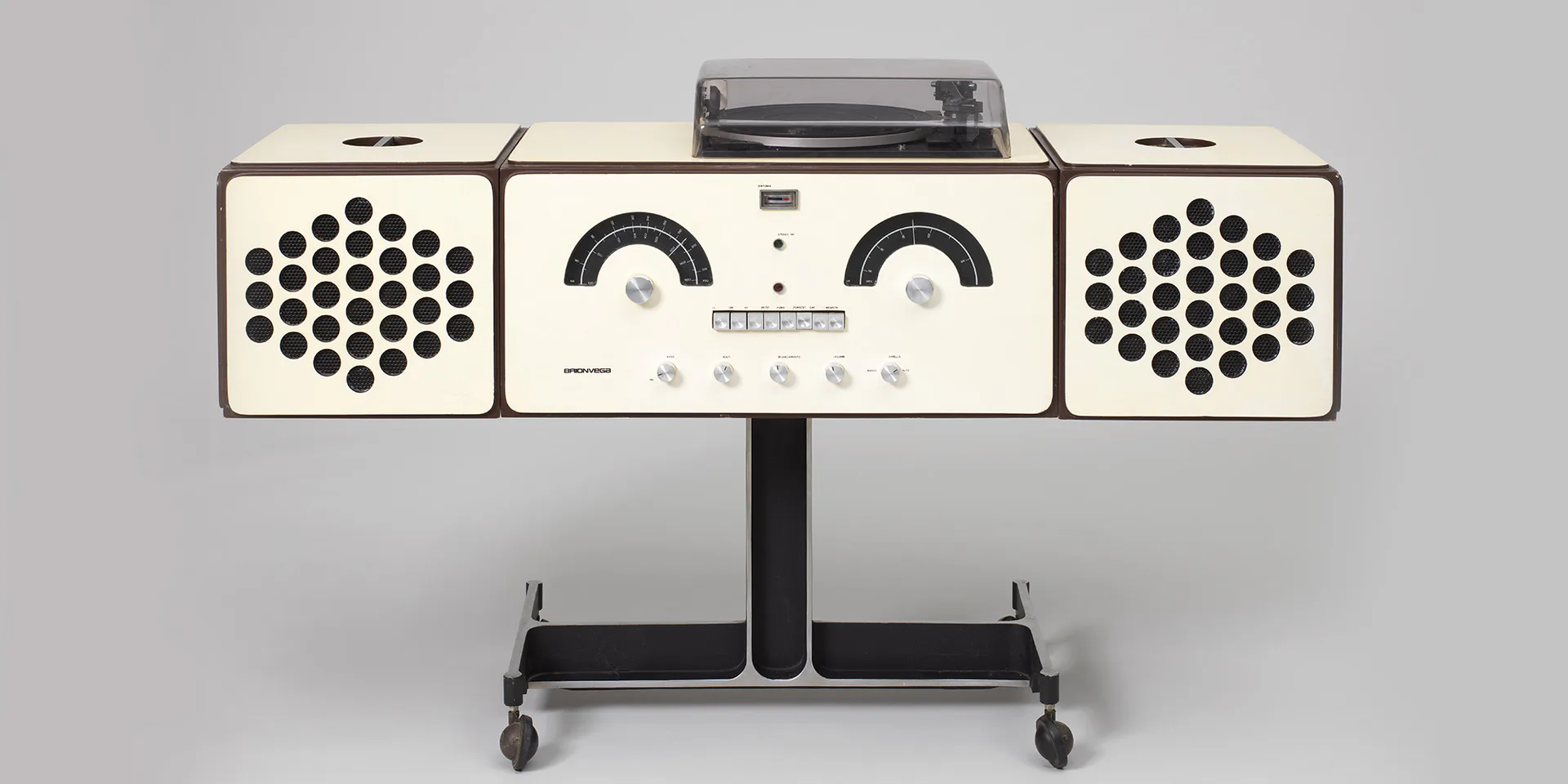 Modernist audio equipment with cream-colored metal housing, twin speaker grilles, central control panel, and turntable mounted on a black stand.