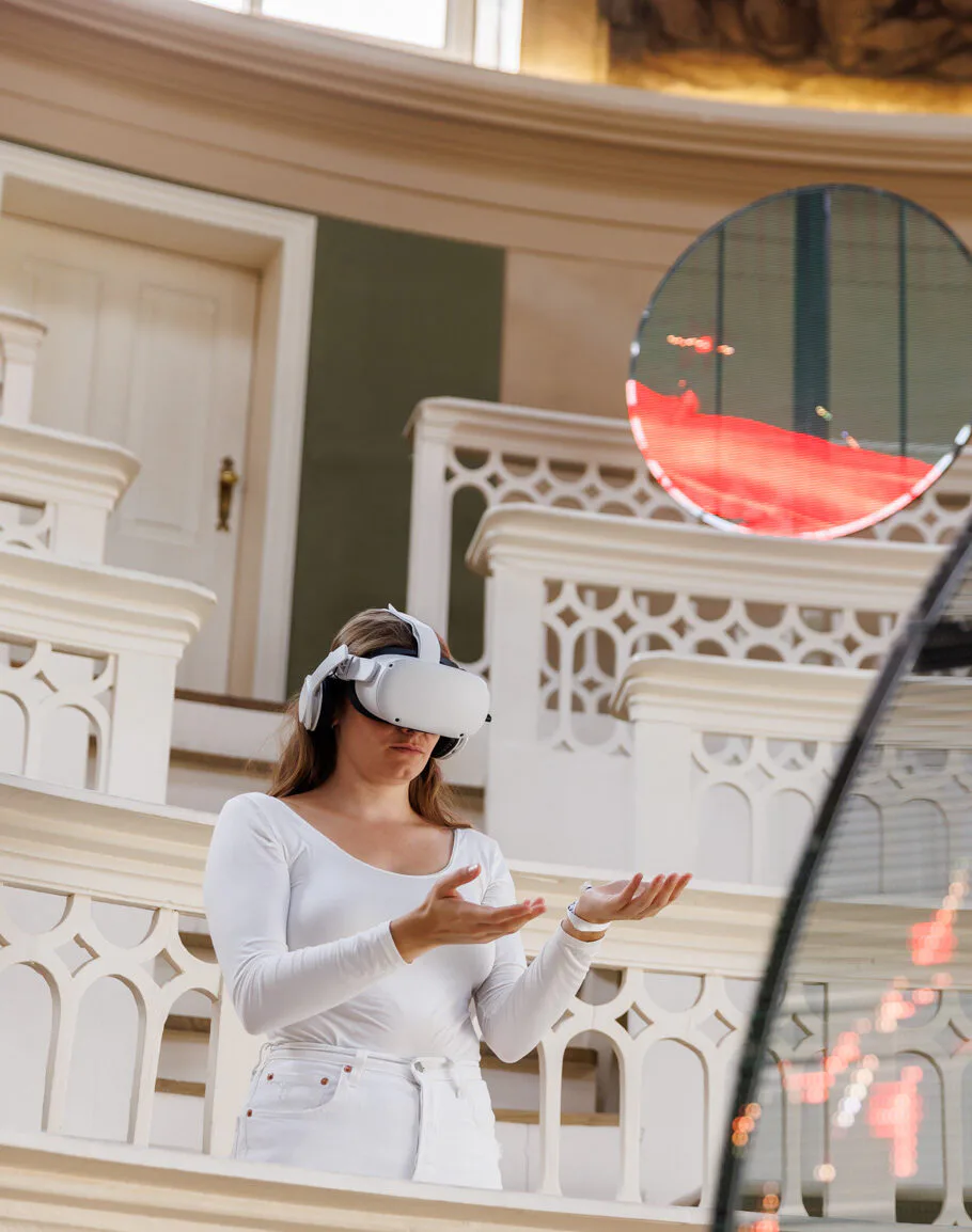 Woman wearing VR headset in ornate interior space with white decorative molding and arched doorways; red digital graphics visible on mirror.