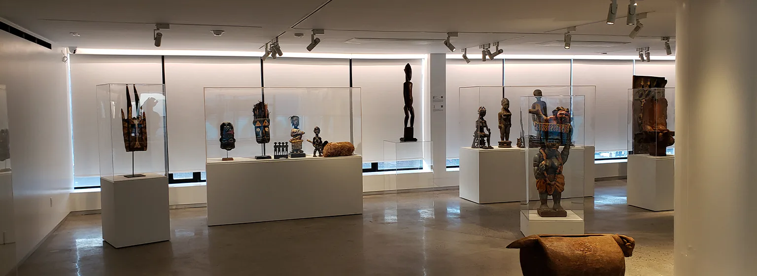 Modern gallery interior with white pedestals displaying African wooden sculptures under spotlights.