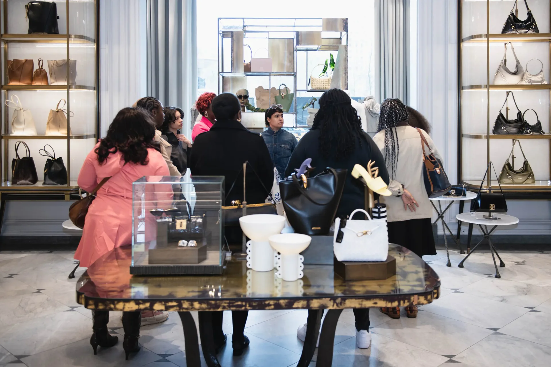 Gallery interior with polished marble floors, wall-mounted shelving displaying handbags, and visitors gathered around a central table.