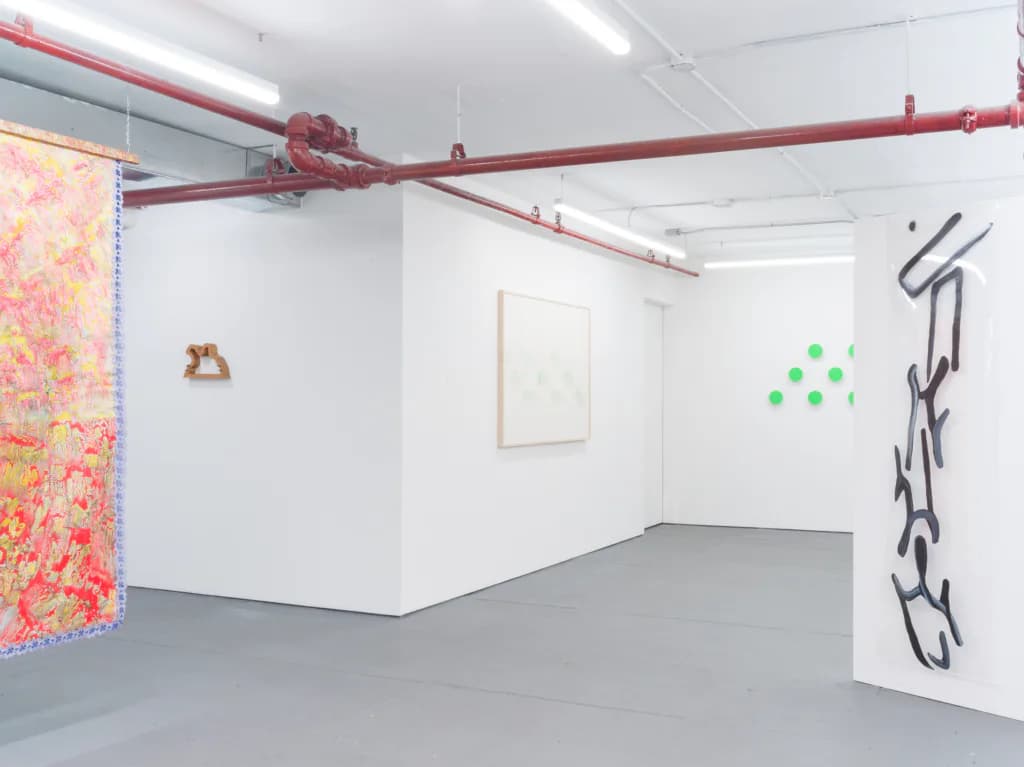 D. D. D. D. gallery interior showing large coral-and-yellow patterned hanging textile, vertical ink drawing on white column, green dot installation, and framed artworks on white walls.