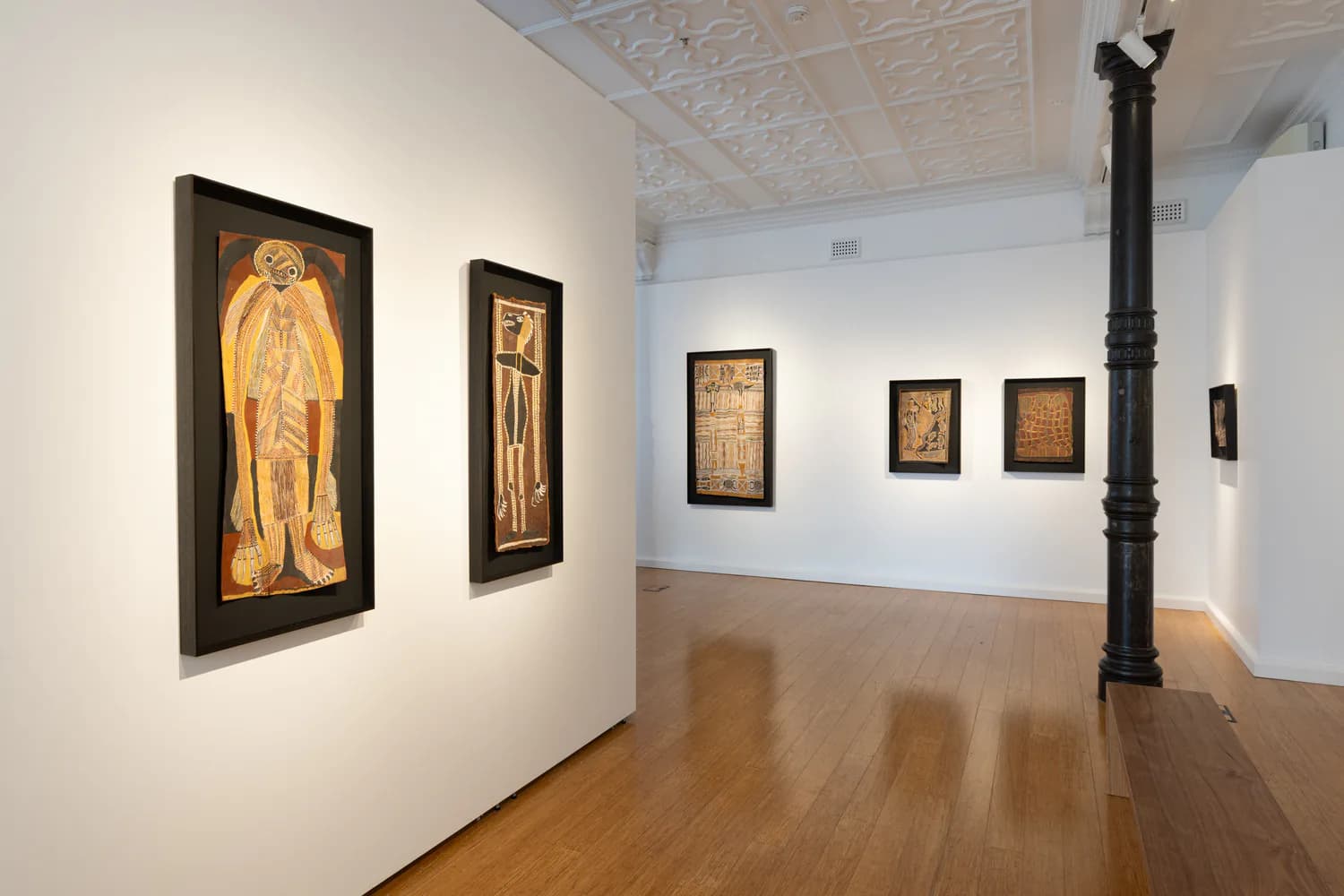 Gallery interior at D Lan Galleries with white walls displaying framed artworks in varying sizes, polished wooden flooring, and a dark sculptural element visible.
