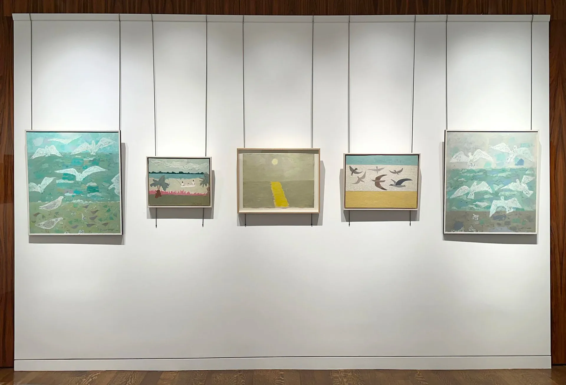 Gallery wall display of five landscape paintings featuring water, trees, and birds in muted greens and blues, suspended at varying heights.