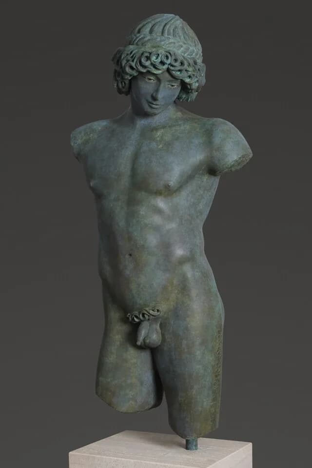 Patinated bronze sculptural torso of a classical male figure with curled hair and drapery details, displayed on a light base against neutral background.