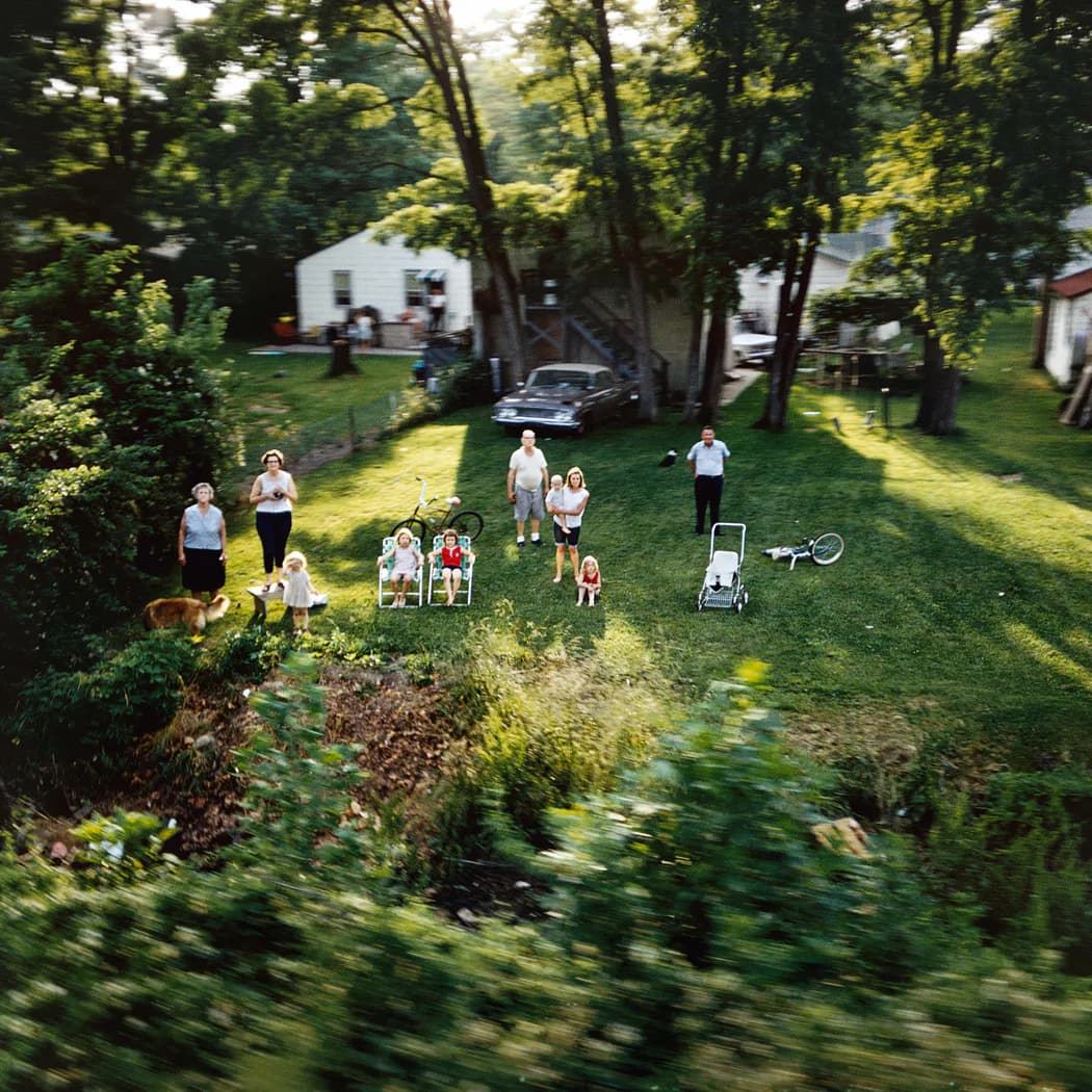 Aerial view of a multigenerational family gathering on a sunlit lawn surrounded by tall trees, with a house and parked car visible in the background.