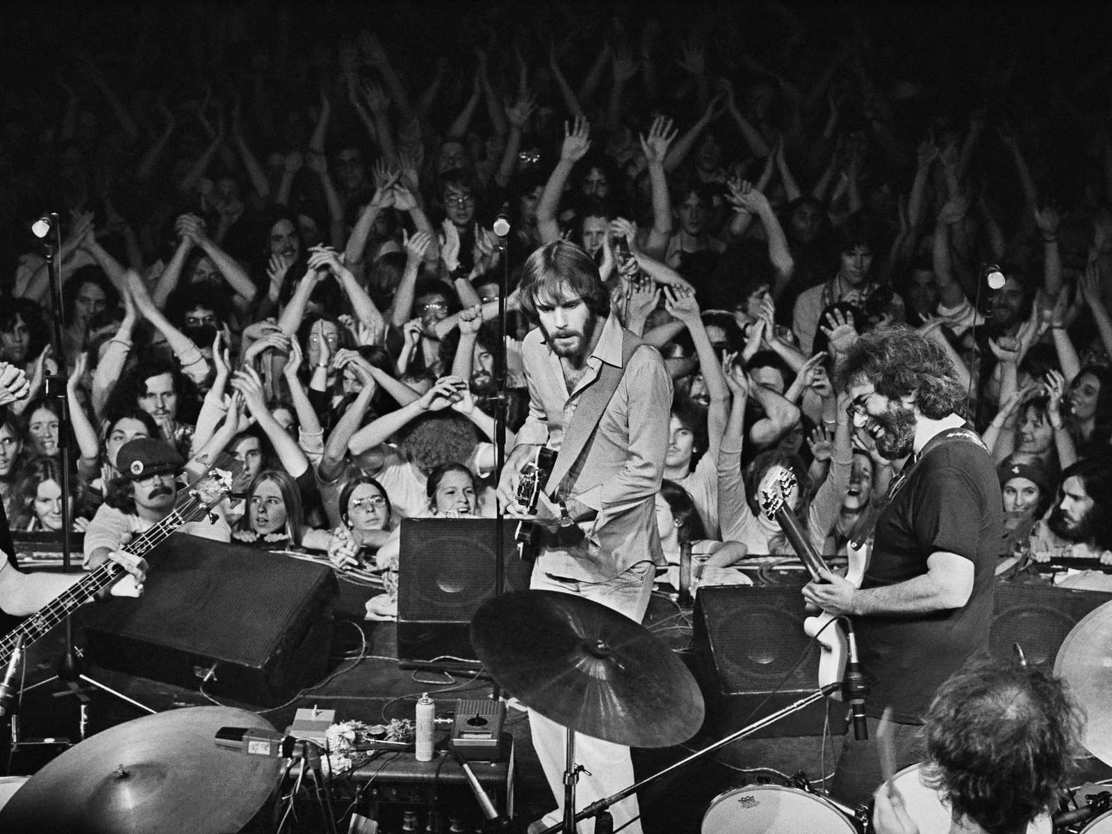 Black-and-white photograph of a band performing on stage with an energetic crowd with raised arms in the background.