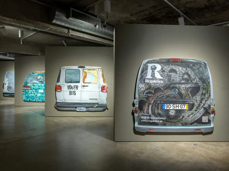 Gallery interior with concrete floor displaying three white vans with various artwork and text on their exteriors as installed sculptures.