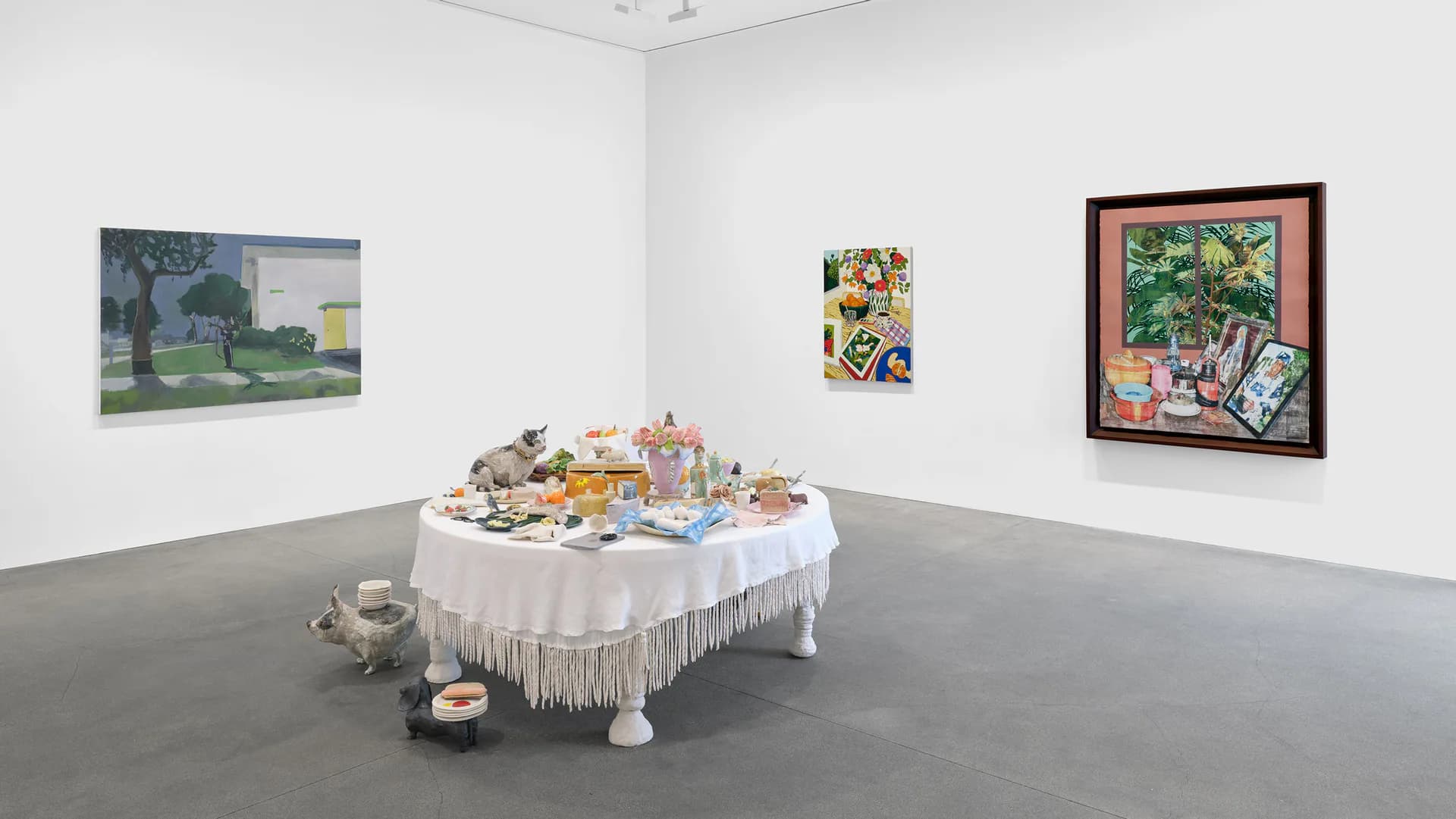 Gallery space with gray floor and white walls. A round white table draped with fringe holds sculptural objects in the center; paintings and mixed-media works hang on walls, including a large landscape and colorful framed pieces.