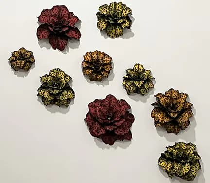 Installation of nine metallic sculptural flowers in gold and deep red, arranged on white wall.