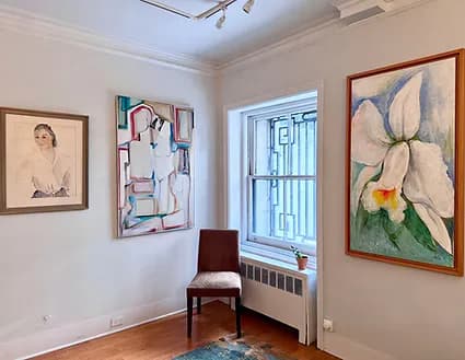 White gallery interior with framed artworks on walls, including abstract painting, figure drawing, and large flower painting; window and chair visible.