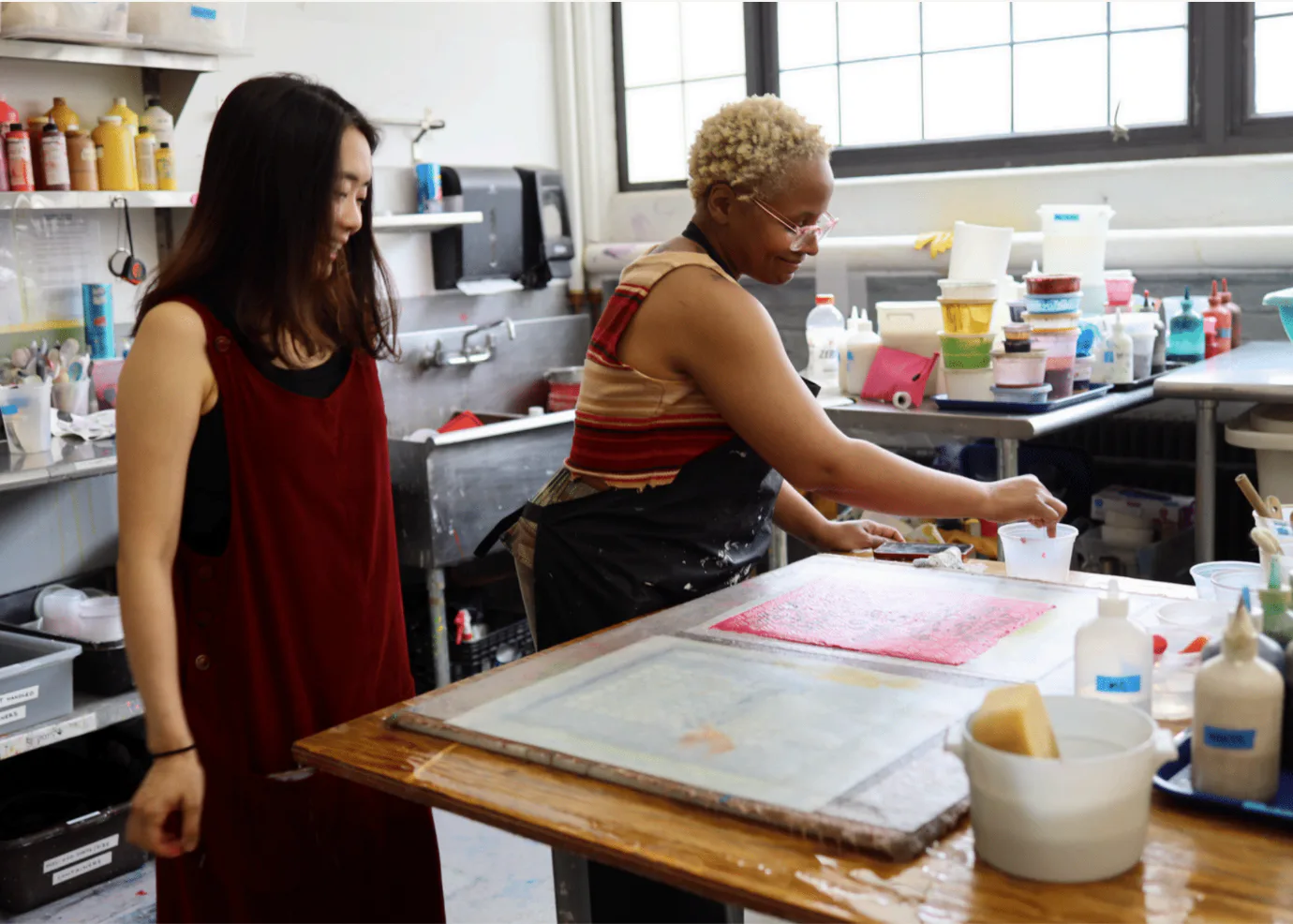 Two artists working at a table in a studio interior with shelves of supplies, natural light from windows, and paper or textile materials in progress.