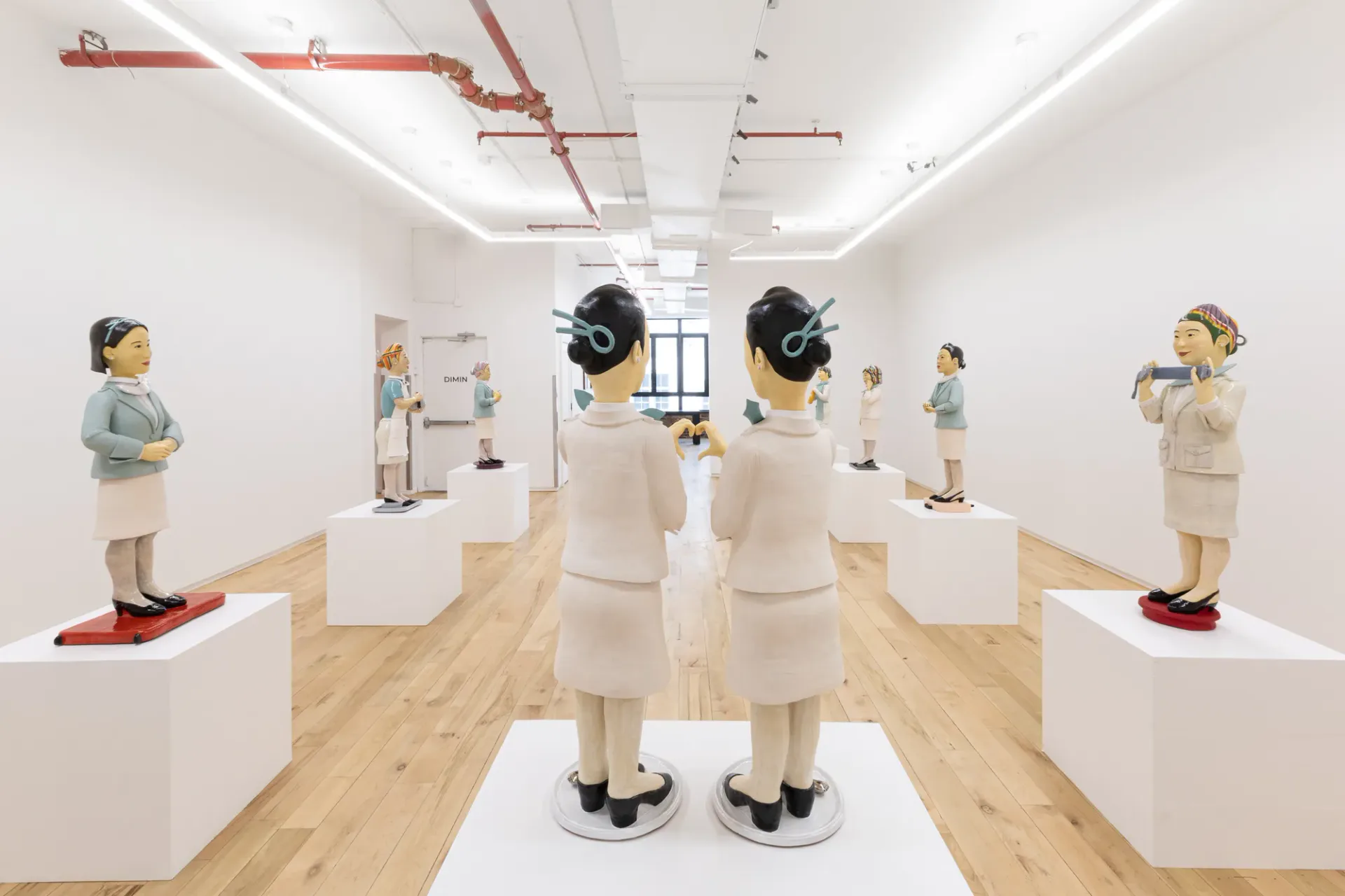 White minimalist gallery interior with wooden floor, displaying ceramic figurative sculptures on white pedestals under linear ceiling lights.