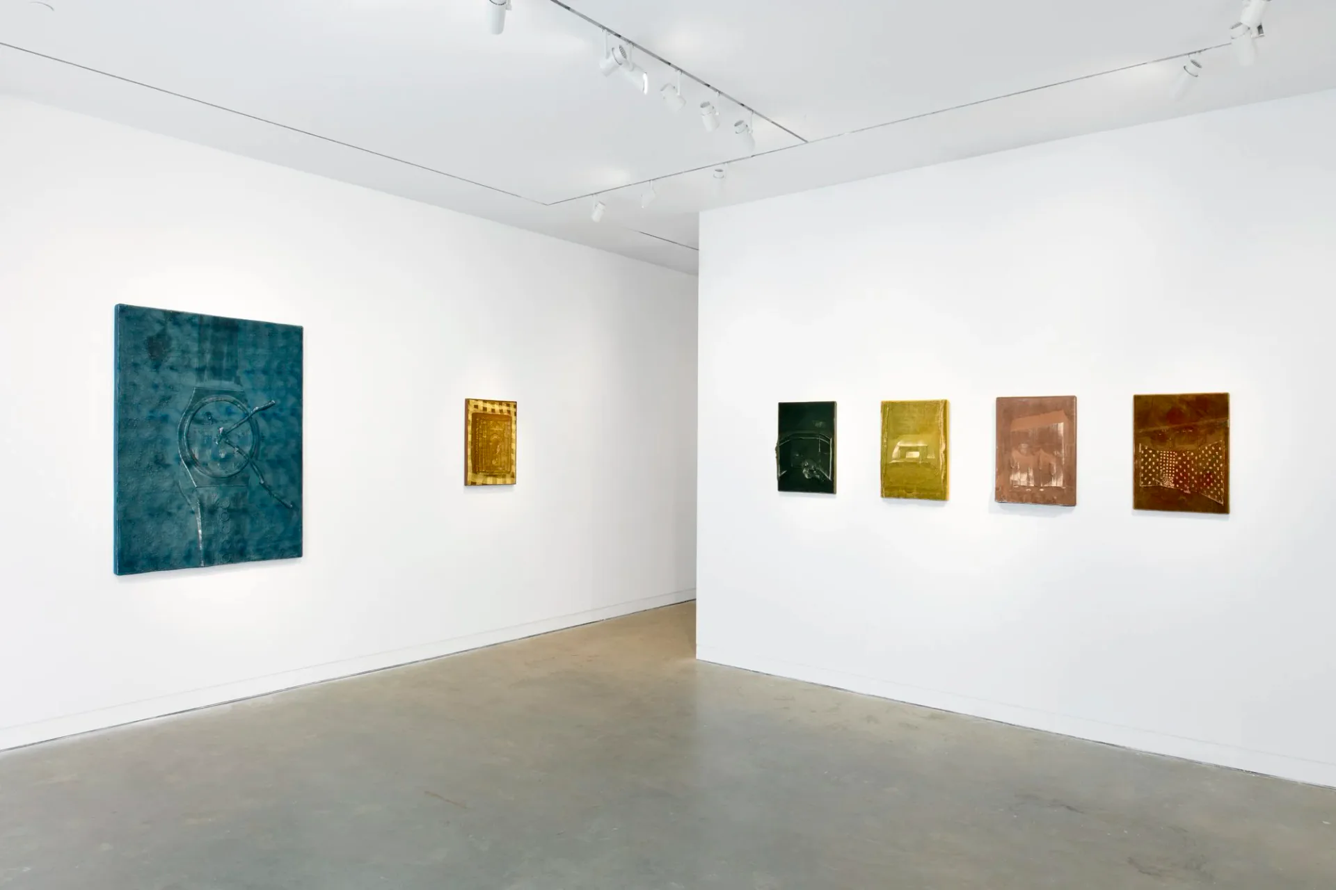 White gallery interior with polished concrete floor displaying abstract artworks: a large teal-blue square on left wall, small golden rectangle in center, and four smaller colorful metallic-toned panels on right wall.