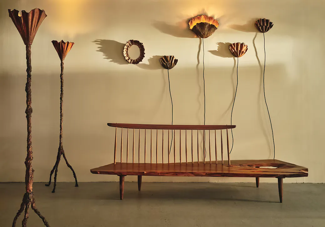 Mixed-media sculptural installation with wooden bench featuring vertical spindle back, standing floor lamps with flared tops, and wall-mounted botanical-form pieces in dark wood.