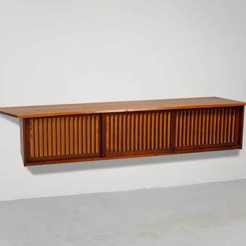 Long wooden credenza or sideboard with vertical slatted cabinet doors in warm cherry or walnut tones, featuring minimal hardware and clean mid-century modern lines.