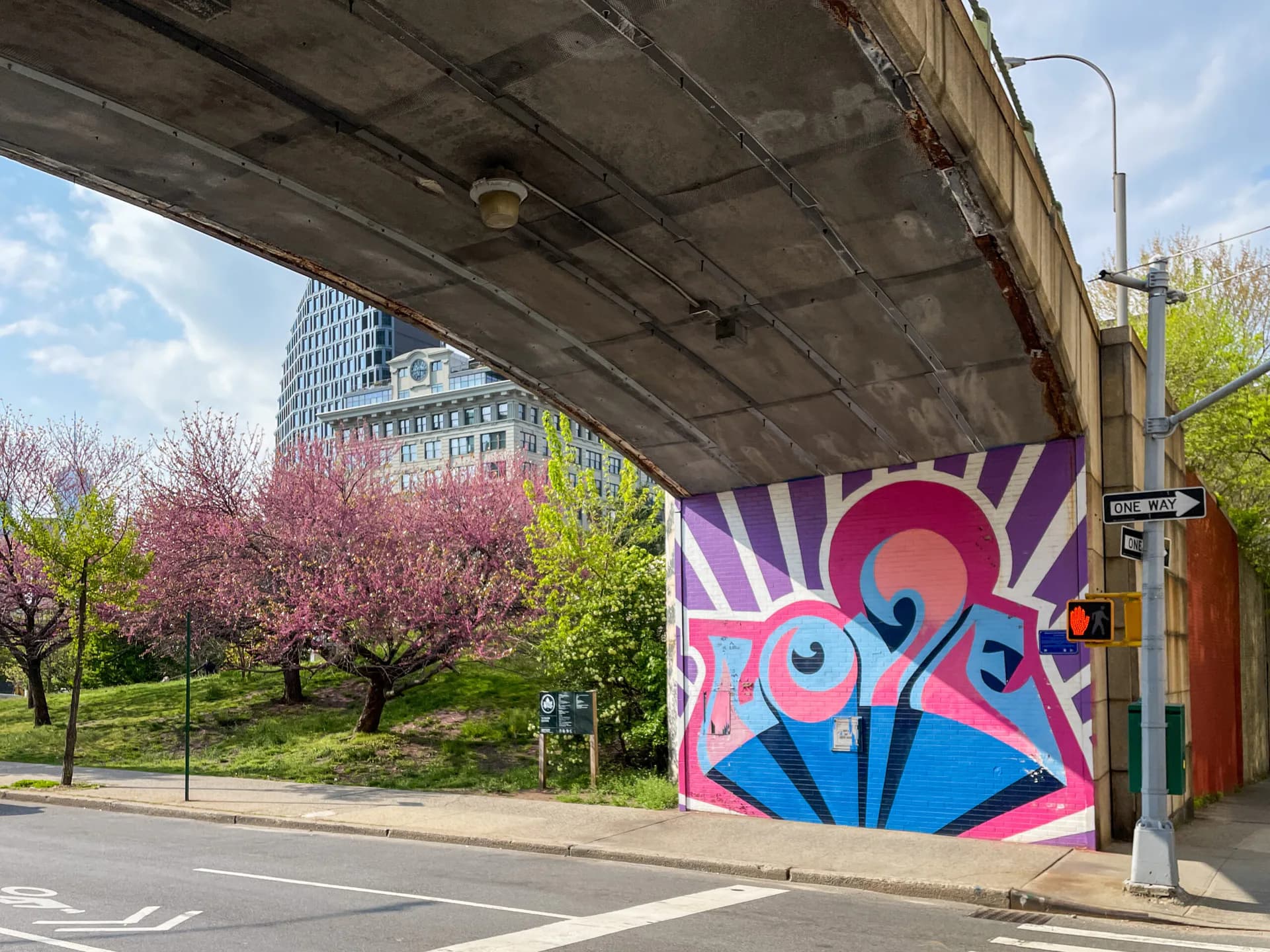 Vibrant street mural under concrete overpass featuring pink, purple, and blue geometric shapes with stylized text and eye-like forms.