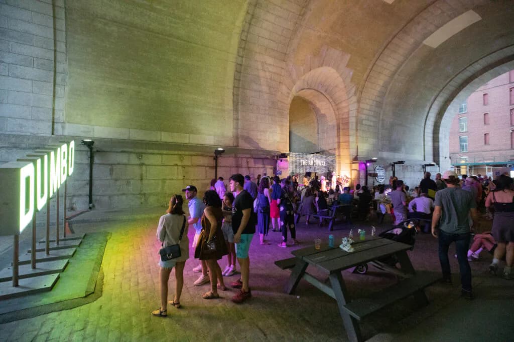 Interior of brick archway venue under Dumbo bridge lit with purple and yellow neon, hosting a gathering with visitors and benches.