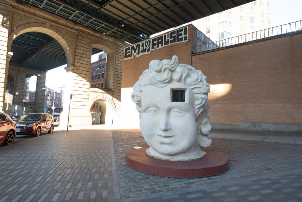 White classical stone head sculpture on red platform beneath Dumbo bridge overpass, with brick buildings and archway visible.