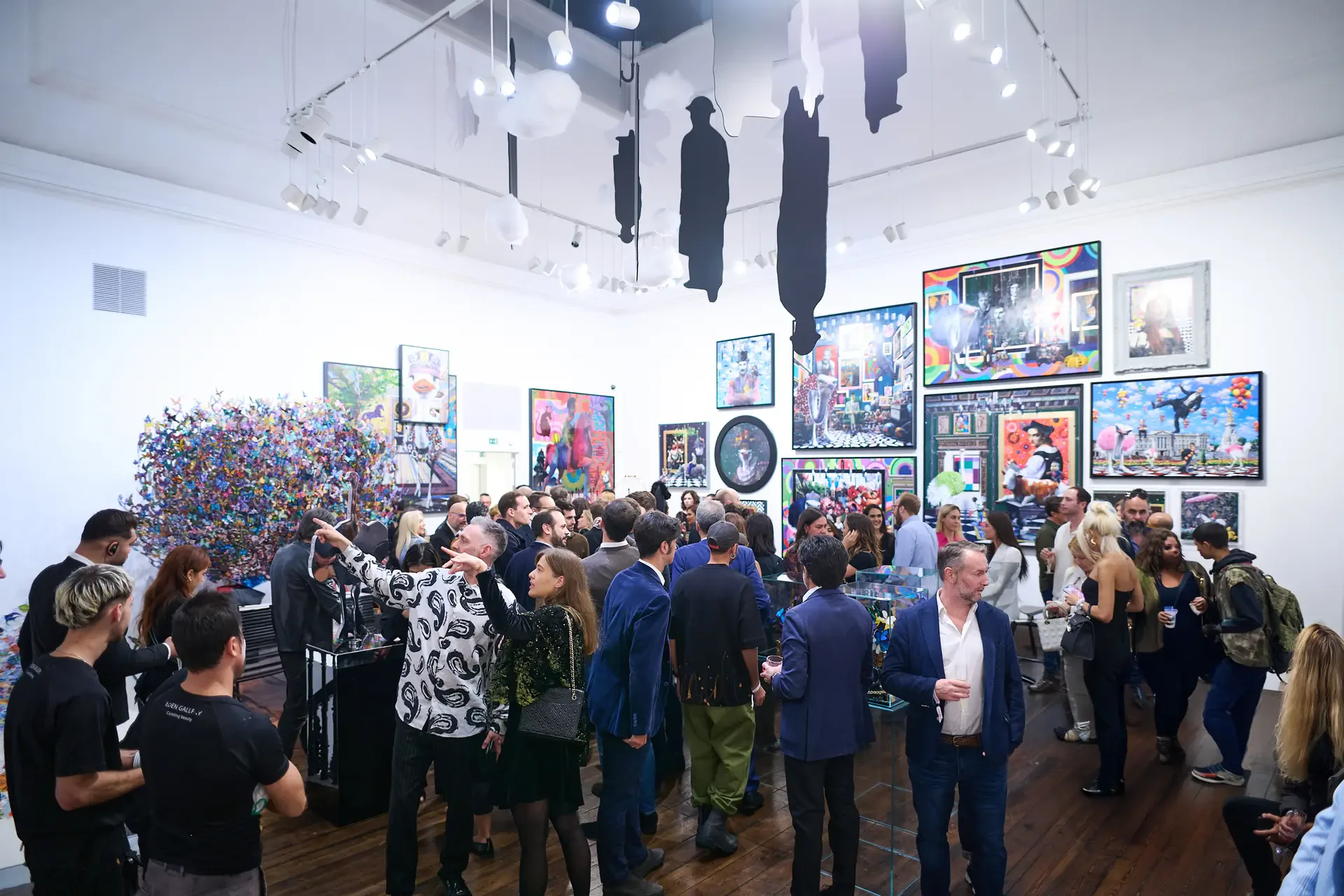 Interior of Eden Gallery with crowded opening reception, white walls displaying colorful artworks, hanging sculptures overhead.