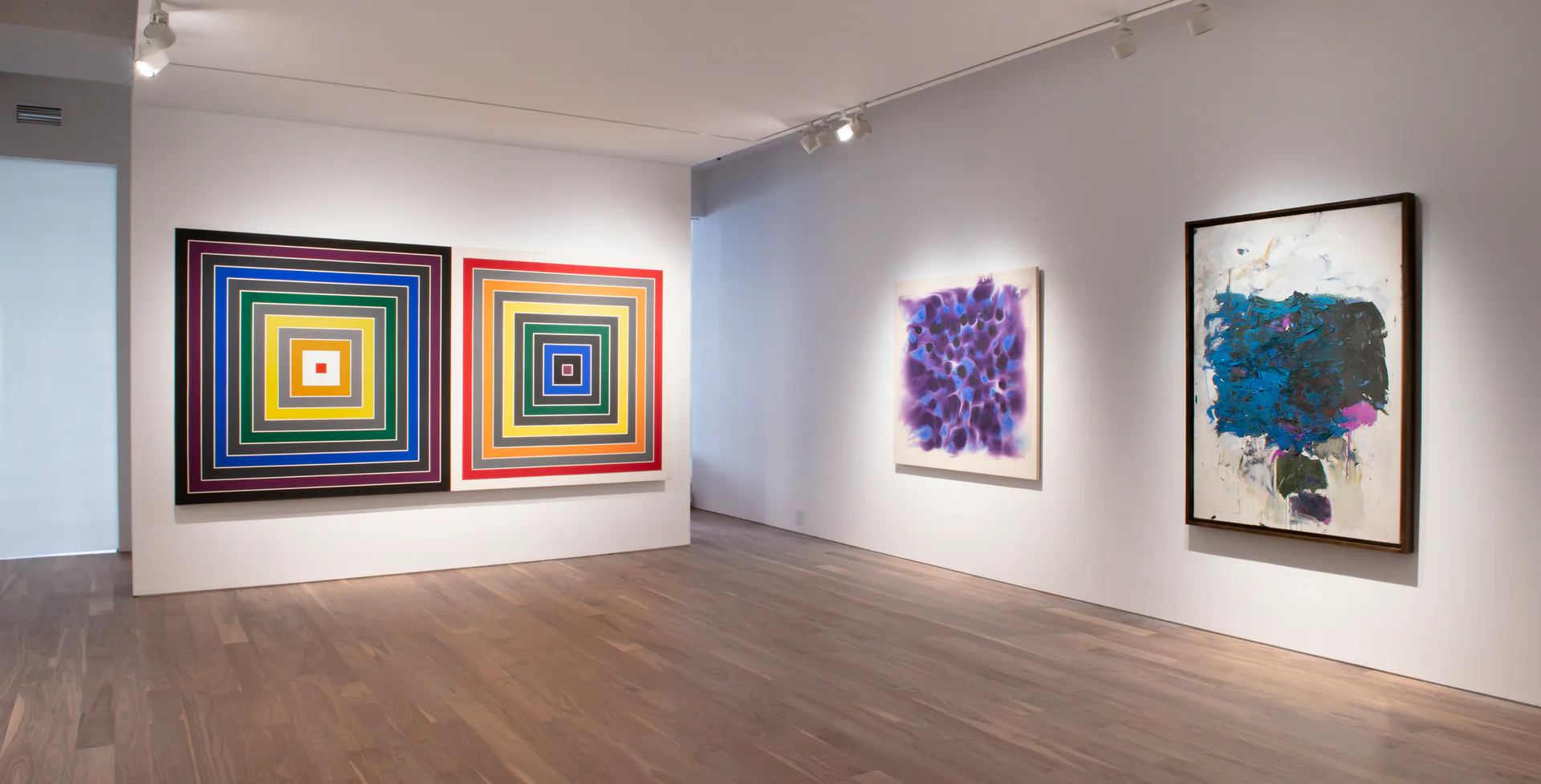 Gallery room with wood floors displaying concentric square paintings in bright colors on left wall, purple and teal abstract works on right walls.